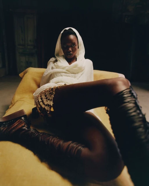Woman with dark skin poses on a yellow cushion wearing tall black boots, a shiny skirt, and a white headscarf in dark indoor lighting.