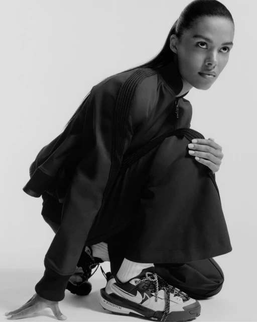 Woman in athletic wear poses in photo studio. She is crouching with Nike sneakers, black jacket, and pants, against a white background.