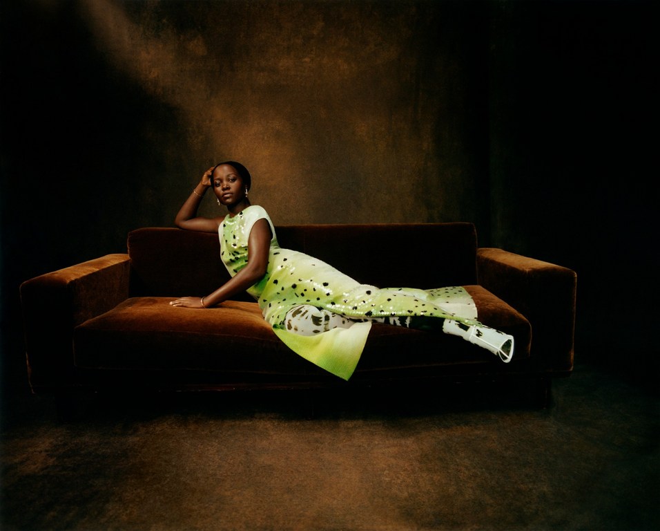 A fashionable black woman with an elegant updo poses on a brown velvet sofa wearing a custom green and black dress in a studio setting.