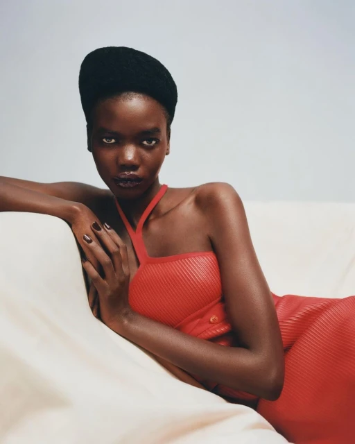 Portrait of a young Black woman lying on a cream-colored sofa. She wears a red, ribbed dress and a black cap while looking at the camera.