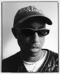 Portrait of Pharrell Williams wearing sunglasses, baseball cap, and diamond necklace photographed by Joshua Woods.