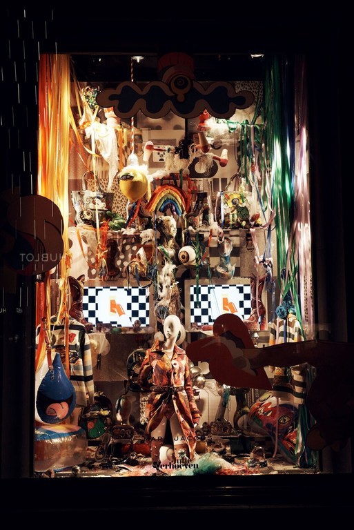Store window display featuring a mannequin in a patterned coat amid an array of colorful objects, screens with checkered patterns, and hanging decorations.