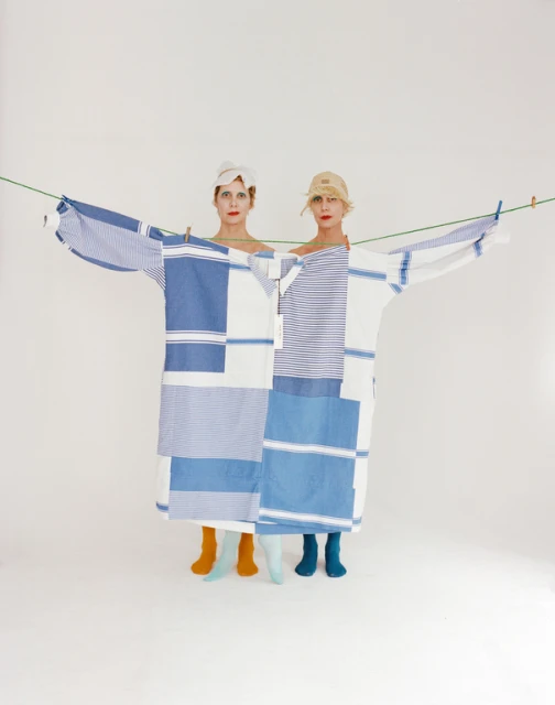 Two women wear a large, shared blue-and-white patchwork shirt on a clothesline against a white backdrop, their colorful socks peeking from underneath.