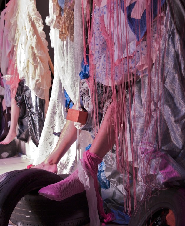 Abstract fashion display featuring a human leg wearing a pink stocking, juxtaposed with layers of fabric, string, and tires.