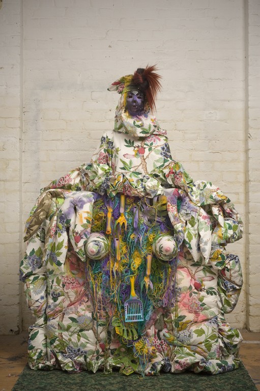 Person in an elaborate, floral-patterned dress and headdress stands indoors with an expressive pose, adorned with garden tools.