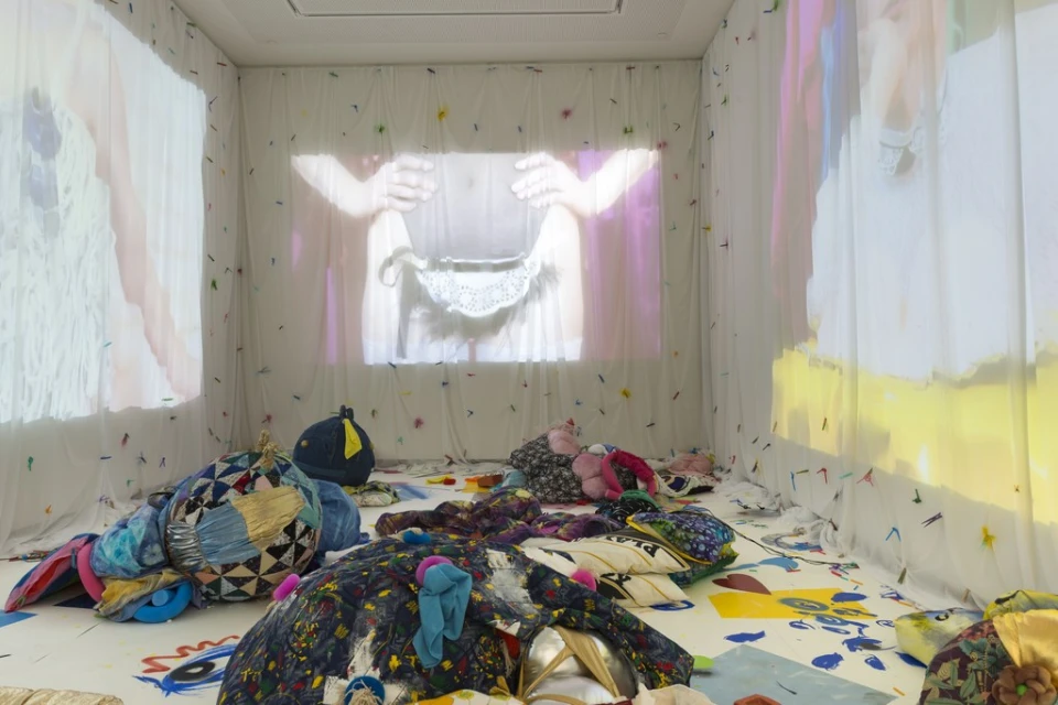 Art installation in a white room shows projections of human torsos on draped white walls and patterned fabric cushions and flooring.