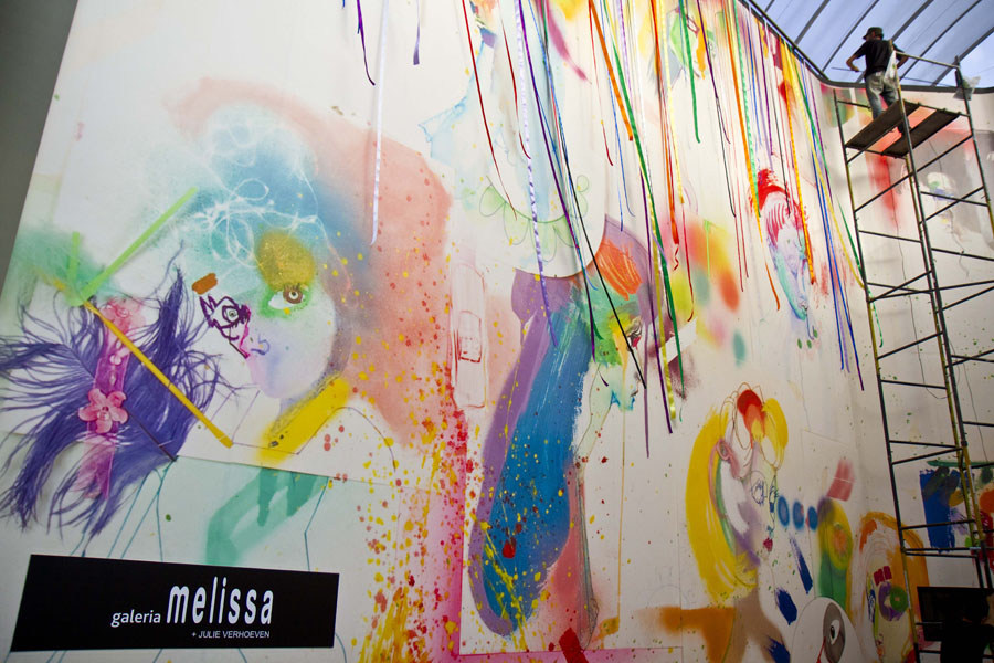Large-scale artwork being created in a bright studio, featuring colorful graffiti-style portraits and an artist on a scaffolding platform.