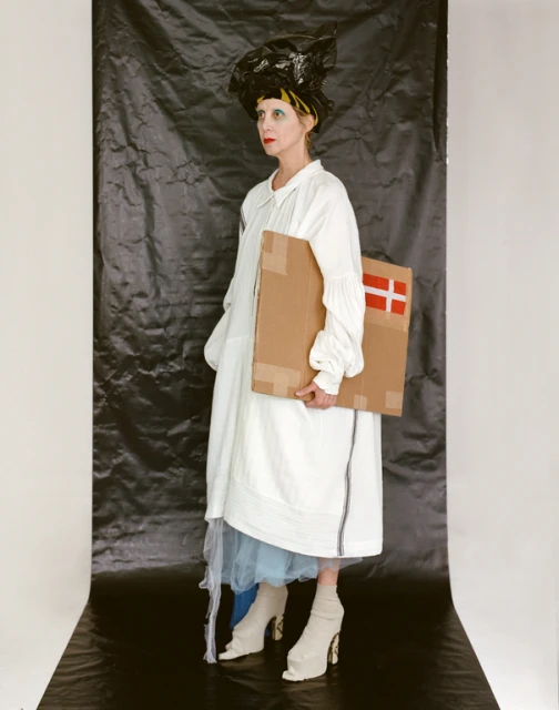 Woman in white dress and headpiece holds a cardboard box with a Danish flag in a photo studio with a black backdrop.