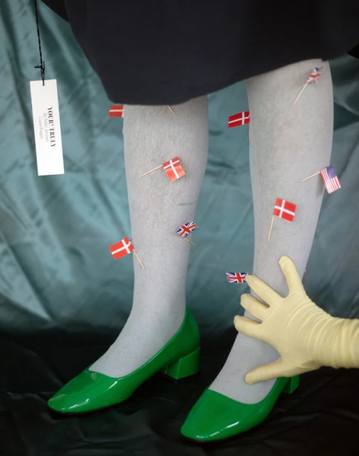 Legs in gray tights and green heels decorated with miniature Danish, British, and American flags held by a hand in a yellow glove.