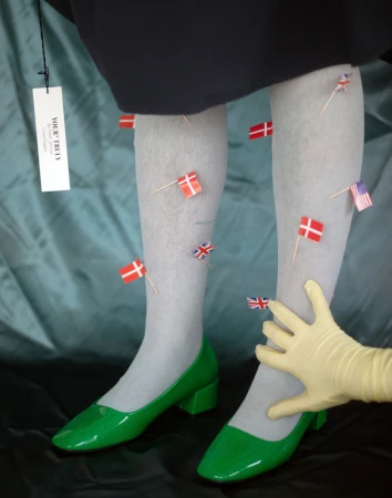 Legs in gray tights and green heels decorated with miniature Danish, British, and American flags held by a hand in a yellow glove.