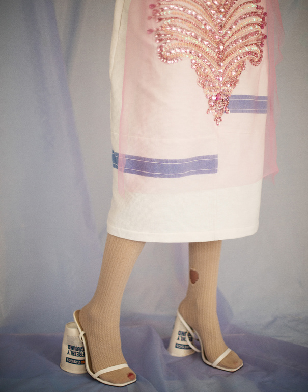 A person's lower half is shown wearing white heels with coffee cup heels, beige socks, and a white dress with pink accents.