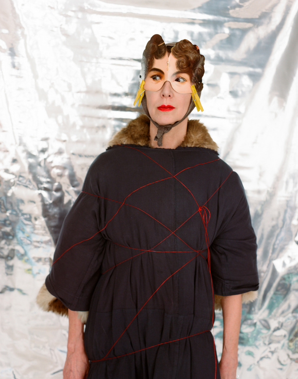 Unique studio shot of a woman wearing a fur-trimmed coat, a headpiece with glasses, and red lipstick against a silver backdrop.