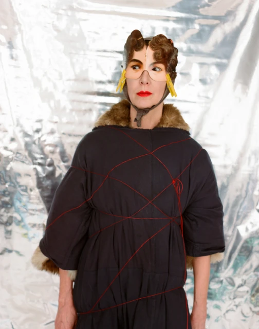 Unique studio shot of a woman wearing a fur-trimmed coat, a headpiece with glasses, and red lipstick against a silver backdrop.