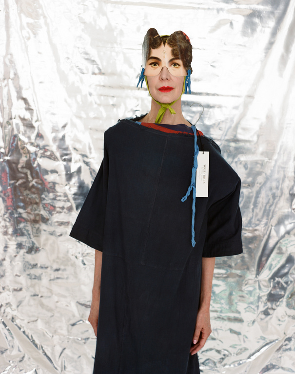 Woman with paper headwear in a dark, oversized dress against a crinkled silver backdrop, showcasing avant-garde fashion and creative styling.