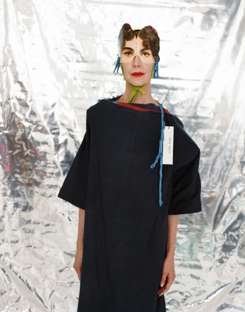 Woman with paper headwear in a dark, oversized dress against a crinkled silver backdrop, showcasing avant-garde fashion and creative styling.
