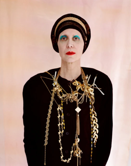 Woman in turban and dark sweater wearing multiple gold necklaces, standing in a studio with a light pink background, portrait orientation.