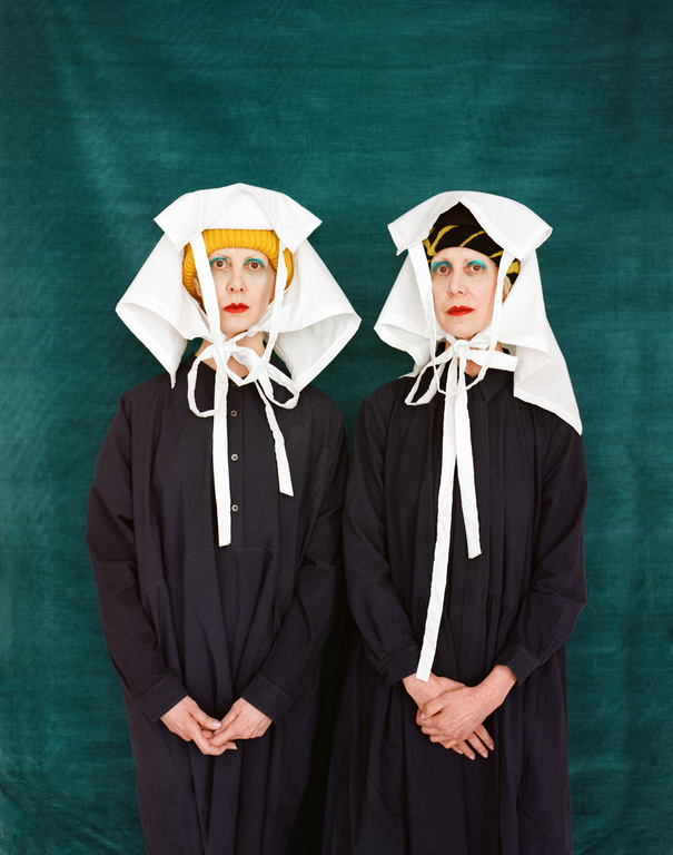 Two women stand in black dresses and white head coverings against a green backdrop, showcasing editorial fashion and unique style.
