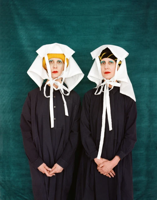 Two women stand in black dresses and white head coverings against a green backdrop, showcasing editorial fashion and unique style.