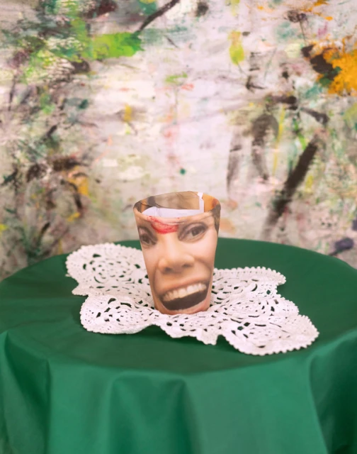 A decorated paper cup with a human face printed on it sits on a green table cloth with a white, decorative doily underneath.