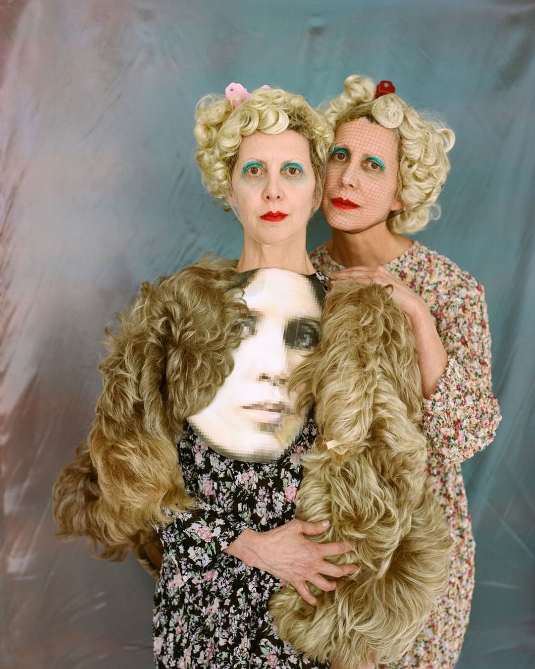 Two adult women with retro-style blonde wigs and makeup posing indoors against a textured blue background, one holding a wigged portrait.
