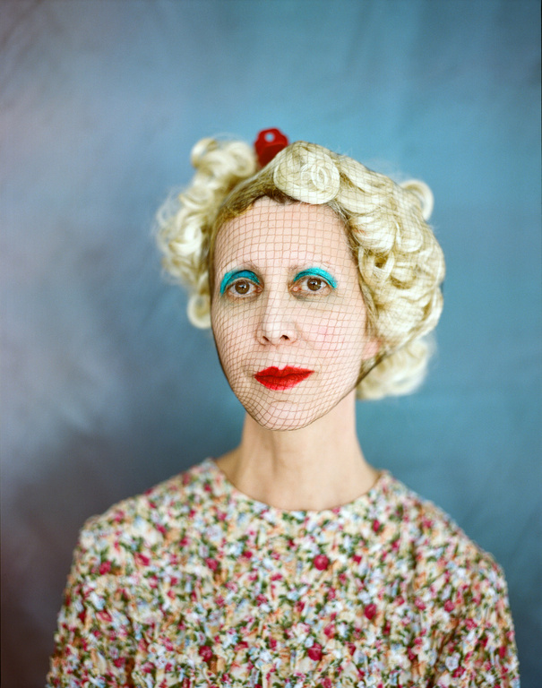 Close-up of a woman with theatrical makeup, a blond wig with a red cherry on top, and a net over her face against a gray background.