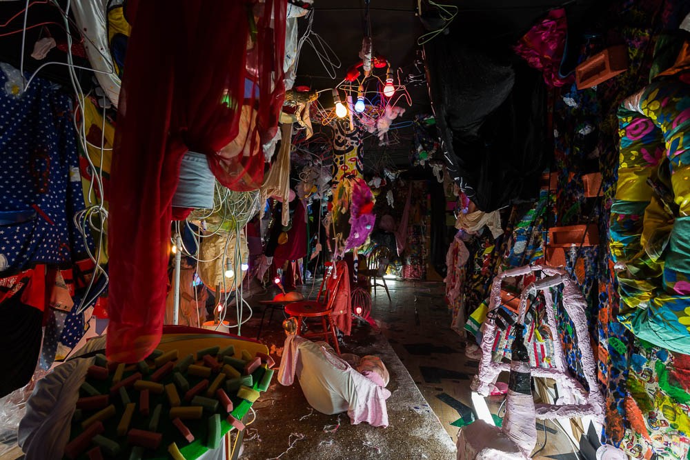 Immersive art installation with colorful, hanging clothes, playful lighting, repurposed furniture and objects creating a unique, textured space.