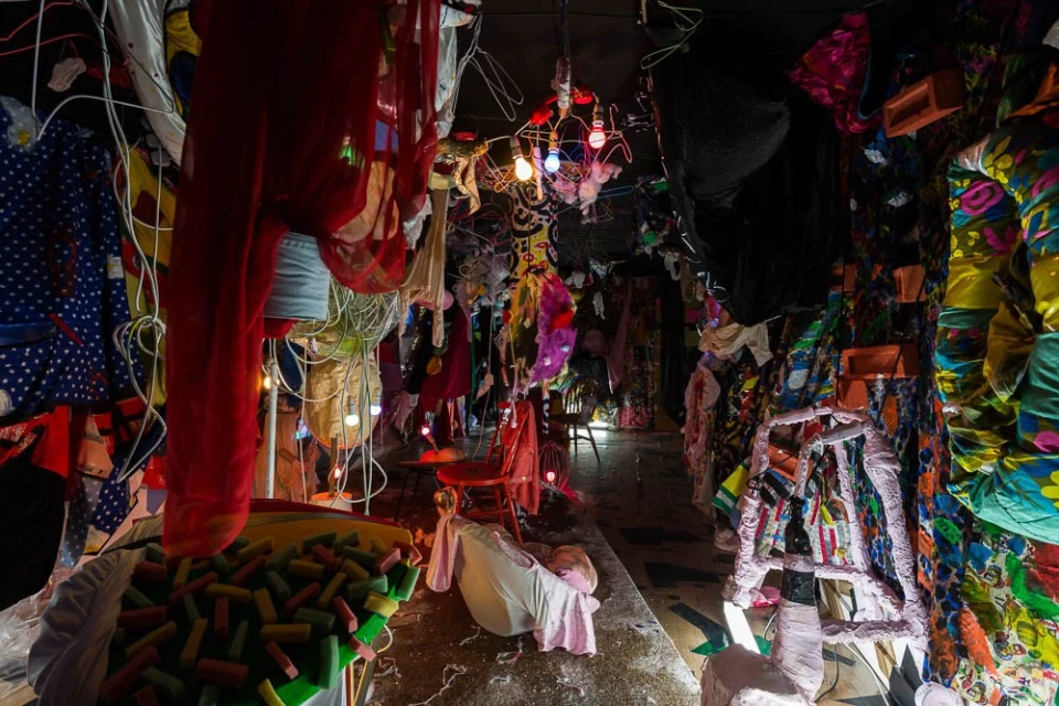 Immersive art installation with colorful, hanging clothes, playful lighting, repurposed furniture and objects creating a unique, textured space.