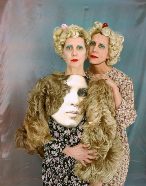 Two women with vintage blonde wigs and floral dresses pose with an antique portrait, creating a surreal and artistic tableau.