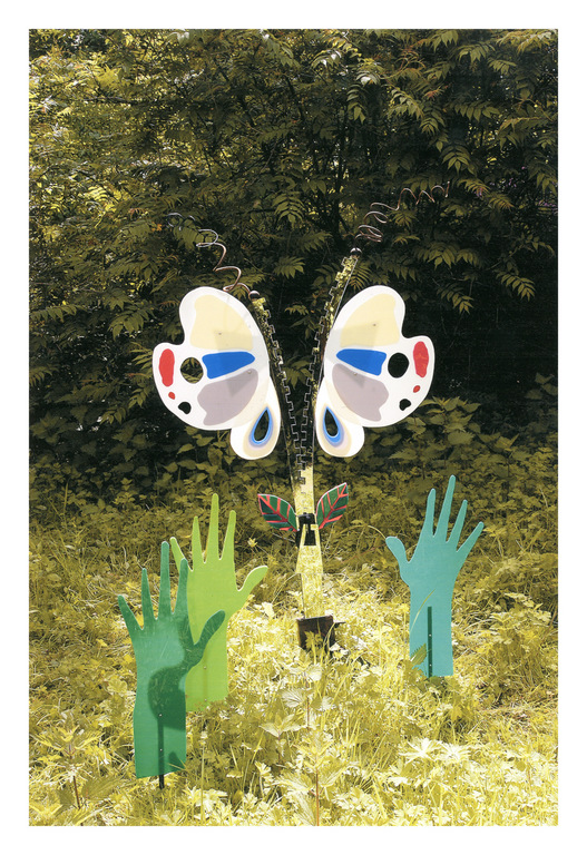Whimsical art installation of butterfly wings and hand-shaped sculptures in a grassy outdoor setting with trees in the background.