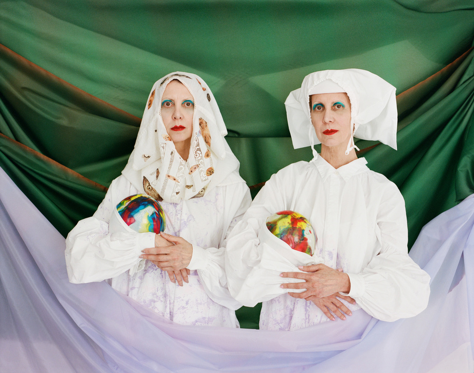 Two women in white religious-style attire hold colorful orbs against a green and lavender draped backdrop in a studio setting.