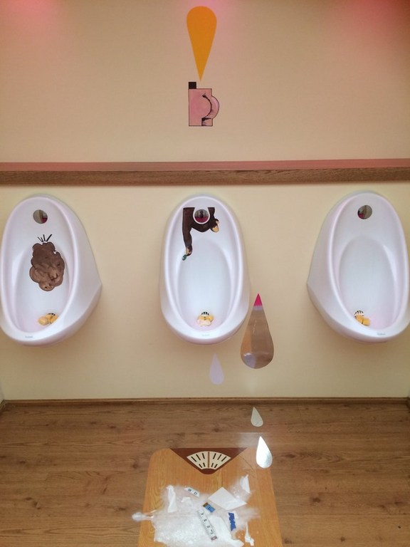 Humorous restroom art featuring urinals with comical characters and illustrations, set against a peach wall and wooden floor.