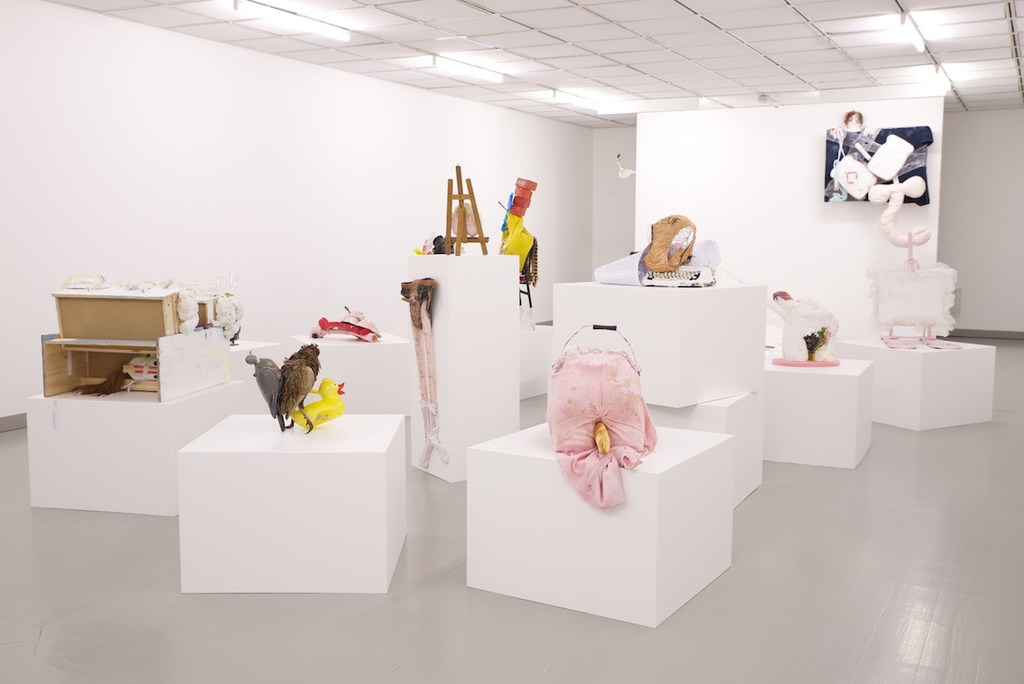 Contemporary art exhibition featuring mixed media sculptures and installations on white cube pedestals in a modern gallery space with white walls.