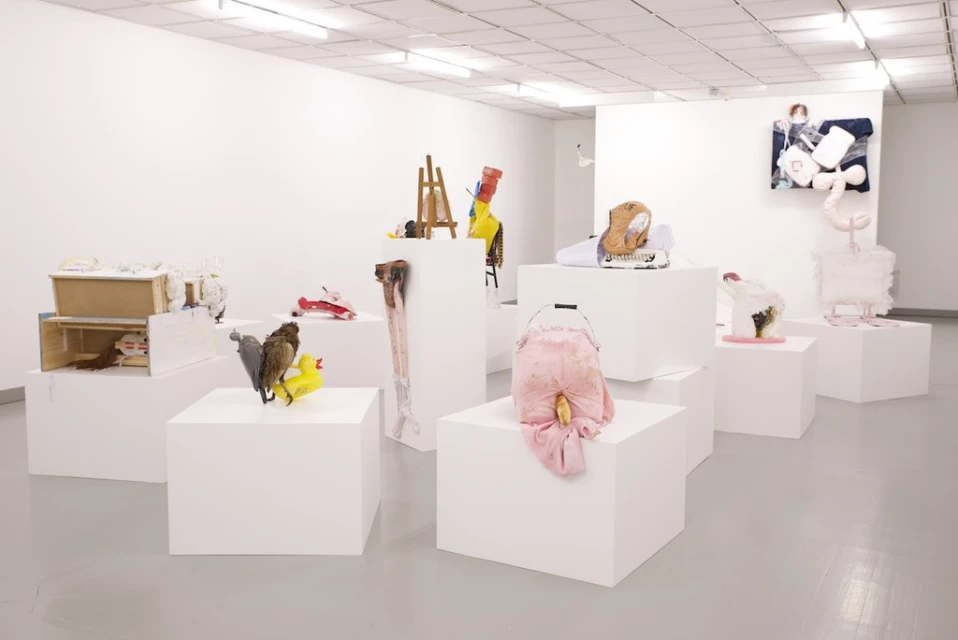 Contemporary art exhibition featuring mixed media sculptures and installations on white cube pedestals in a modern gallery space with white walls.