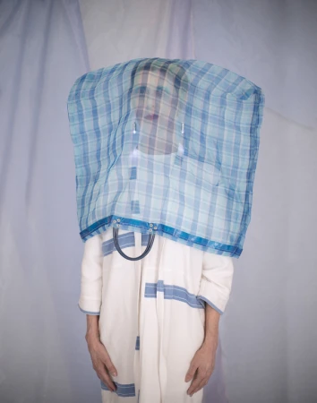 Person standing behind a translucent blue plaid bag covering their head wearing an off-white coat against a pale fabric backdrop
