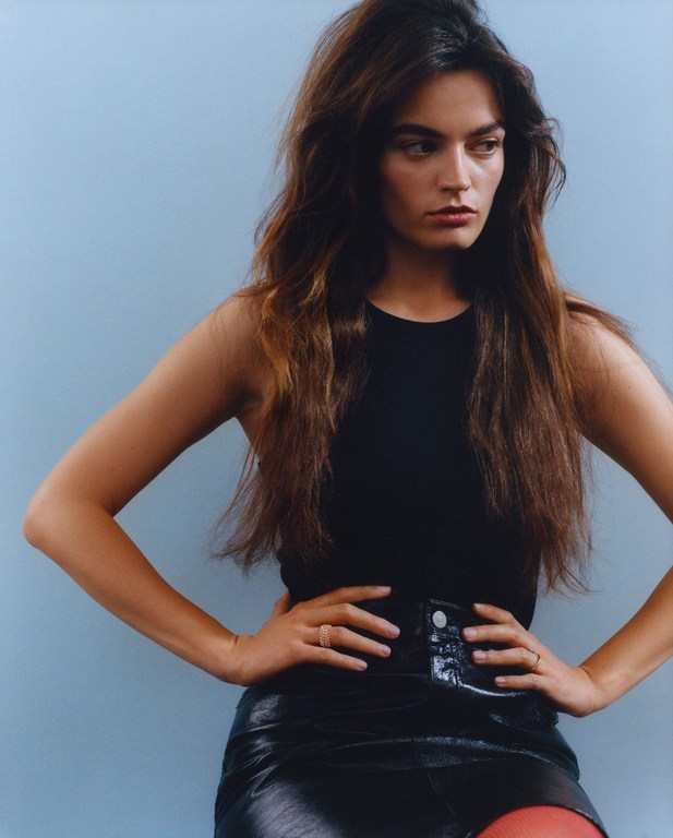 Portrait of a woman with long brunette hair wearing a black top and leather skirt, posing in front of a light blue background.