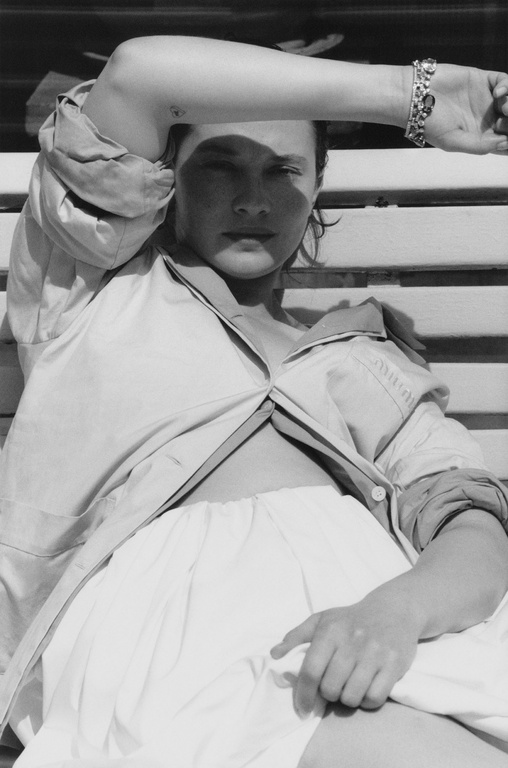 A black and white portrait of a woman reclining, wearing an unbuttoned collared shirt and skirt, with a bracelet on her wrist.