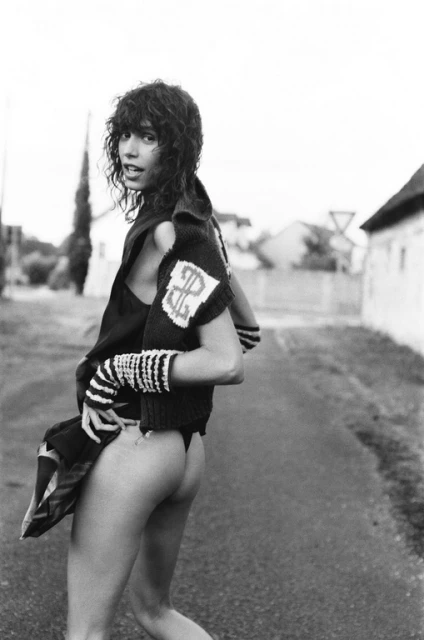 Monochrome outdoor shot of a woman with curly hair wearing a sweater and posing on a road with residential buildings in the background.