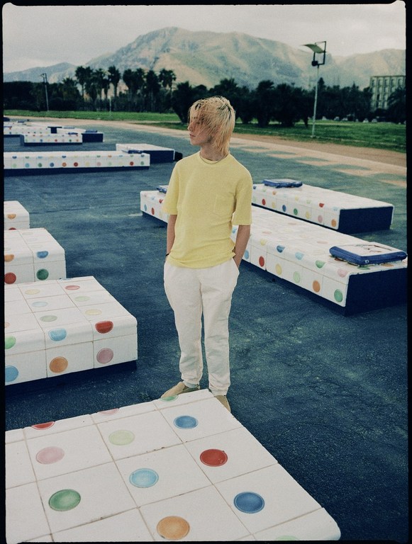 Outdoor portrait of a person with blond hair wearing a yellow t-shirt and white pants, standing near colorful benches and a distant mountain.
