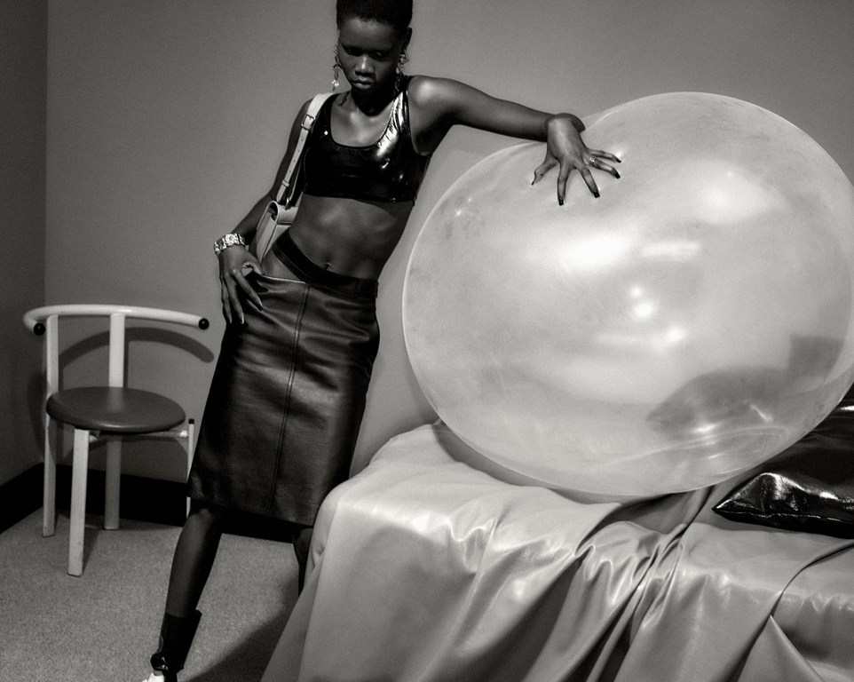 Monochrome portrait of a stylish woman in a skirt and shiny top, leaning against a large transparent bubble with a chair in the background.