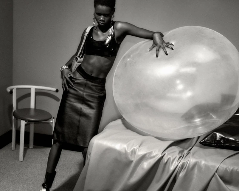Monochrome portrait of a stylish woman in a skirt and shiny top, leaning against a large transparent bubble with a chair in the background.