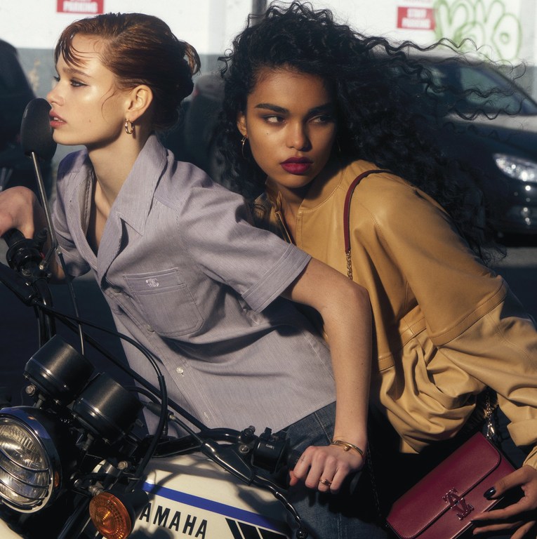 Two fashion models pose on a Yamaha motorcycle in an urban setting with neutral tones and soft lighting, showcasing contemporary style.