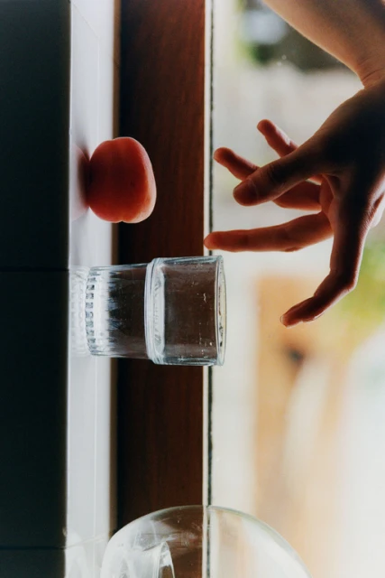 Still life photograph of a peach, drinking glasses, and a human hand with outstretched fingers in a brightly lit indoor setting.