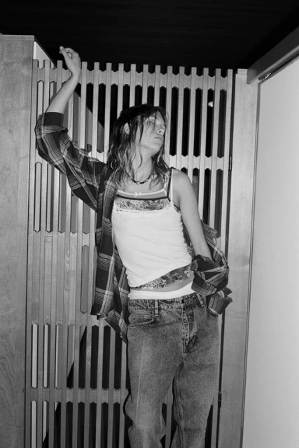 Monochrome portrait of a young adult woman dancing indoors wearing a white tank top, blue jeans, and a plaid shirt.
