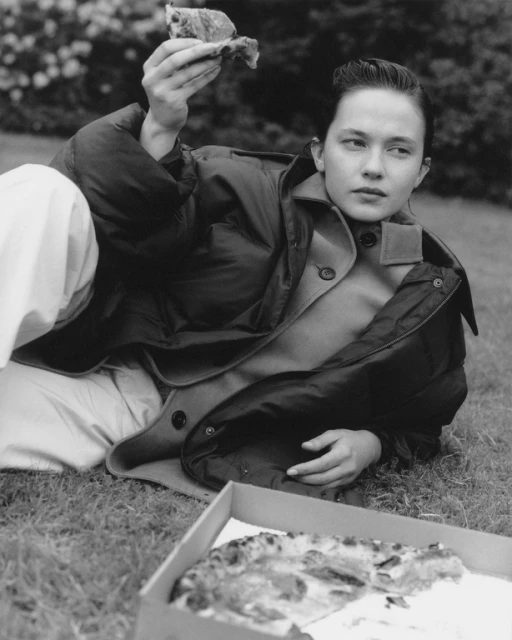Woman in puffer jacket lies on grass with pizza.  Black and white portrait shows the subject holding a slice, near an open pizza box.