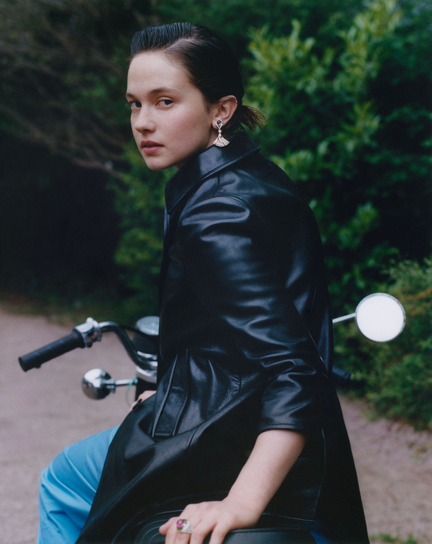 A young woman with dark hair, wearing a black leather jacket and light blue pants, sits on a black motorcycle in a wooded area.