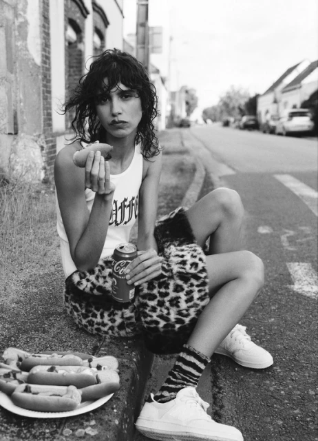 A young adult female model sits on a curb in a street setting, eating a hotdog and holding a soda can, with a plate of hotdogs beside her.