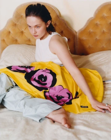 A young woman with wet hair lies on a bed with a yellow graphic-print dress. She is indoors with a beige headboard and white sheets.