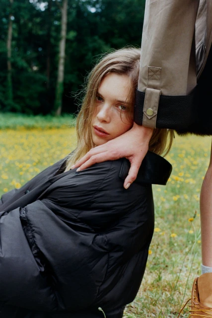 Blonde woman in a black jacket outside in a grassy, green field with yellow flowers, hand on shoulder, and another figure beside her.