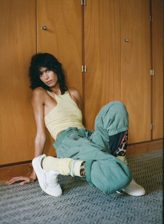 A woman with dark curly hair sits on the floor against wooden paneling, wearing a yellow top, green pants, white sneakers, and yellow socks.