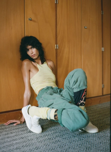 A woman with dark curly hair sits on the floor against wooden paneling, wearing a yellow top, green pants, white sneakers, and yellow socks.